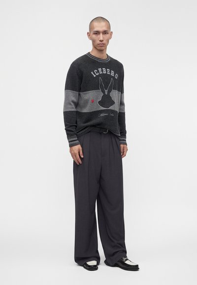Homme portant un pull rayé noir et gris avec un logo de lapin, un pantalon noir large et des chaussures noires et blanches, debout sur un fond blanc.