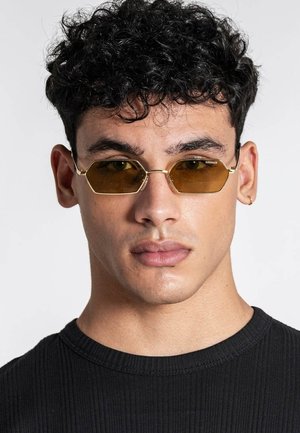 Joven con cabello rizado que lleva gafas de sol hexagonales ámbar con montura dorada y una camiseta negra de canalé, con un pendiente de aro dorado en la oreja izquierda.