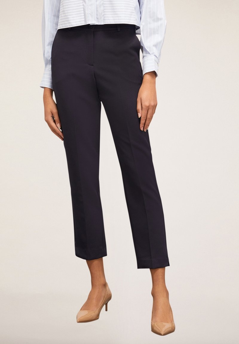 Pantalons courts bleu marine avec une coupe ajustée, devant plat et poches latérales, associés à des escarpins nude sur un fond neutre.