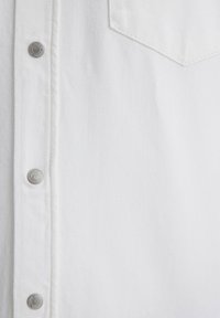 Weiße Denim-Bluse mit Knopfverschluss, einer einzelnen Brusttasche und silberfarbenen Hardware-Details sowie einer strukturierten Stoffoberfläche.