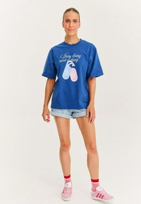 Modrá oversized tričko s bielymi a pastelovo ružovými grafikou textu, skombinované s džínsovými šortkami a ružovými teniskami s červenými akcentmi.