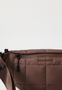 Smeđa pufasta torba preko tijela s strukturovanom površinom, zip zatvaračem, podesivim remenom i utisnutim logotipom. Glatki materijal s suptilnim sjajem.