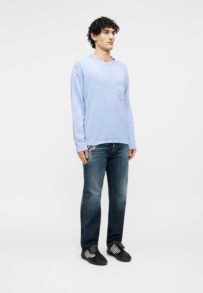 Homme debout portant un pull à manches longues bleu clair avec une poche poitrine, un jean bleu délavé avec une petite déchirure, et des baskets noires à rayures blanches.