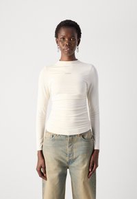 Kremfarget langermet topp med høy hals, med samlet tekstur og subtil merkevare. Kombinert med lysvaskede, løstsittende jeans.