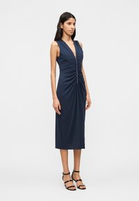 Γυναίκα που στέκεται φορώντας ένα αμάνικο ναυτικό μπλε μίντι φόρεμα με ρίγες και φερμουάρ μπροστά, καθώς και μαύρα σανδάλια με τακούνι και λουράκια, σε λευκό φόντο.