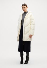 Pildīts krēmkrāsas mētelis ar rāvējslēdzēju priekšā un kapuci, apvienots ar pelēku turtleneck un tumšu svārku. Spīdīgi melni augstpēdu zābaki.