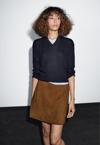 Schwarzer Strickpullover mit V-Ausschnitt und hochgekrempelten Ärmeln, kombiniert mit einem braunen Minirock aus Wildleder. Einfaches, figurbetontes Design und weiche Textur.