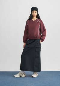 Maroon nylon pullover with white piping and a v-neck, paired with a long black skirt featuring cargo pockets. Worn with beige sneakers.