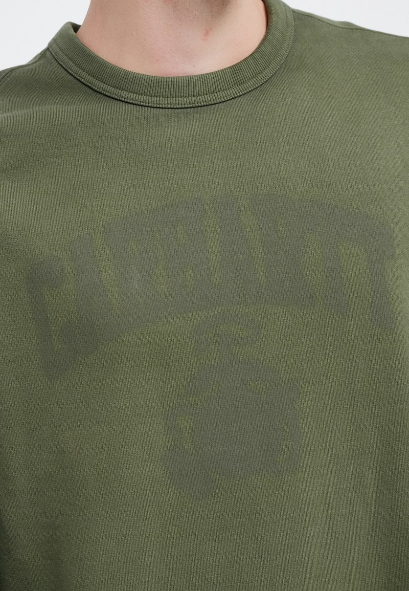 Olive green sweatshirt with a crew neck, featuring a faded logo and graphic print on the front. Made of soft, textured fabric.
