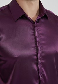 Chemise en satin violet avec une texture lisse, dotée d'un col pointu, d'un devant boutonné et d'un léger éclat. Les détails incluent des boutons assortis.