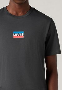 Černé bavlněné tričko s kulatým výstřihem, na přední straně s logem v modré, růžové a bílé barvě v obdélníkovém rámečku.