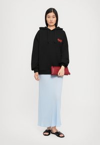 Sweatshirt preto com capuz e texto vermelho, combinado com uma saia maxi justa de tom azul claro. O modelo segura uma bolsa vermelha e usa slides pretos.