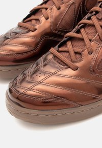 Pruun metallik tennisjalatsid koos tekstureeritud kangaga ja paksude kummitaldadega, millel on detailne õmblus ja ristnostud paelad.