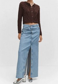 Cardigan en tricot marron avec un motif en losanges, accompagné d'une jupe en denim bleu clair avec une fermeture à boutons et une fente sur le devant. Chaussures argentées à talons blocs.