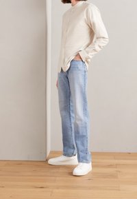 Hellbeige Langarm-Hemd mit Knöpfen und geradem Schnitt, kombiniert mit hellblauen geraden Jeans und weißen Sneakers. Holzboden im Hintergrund.