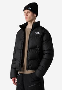 Vīrietis melnā pūkainā jaku un atbilstošu cepuri ar The North Face logotipu, bēša džemperi un melnas bikses, stāv ar rokām kabatās.