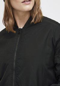 Jachetă bomber neagră cu o textură netedă și mată, dotată cu un fermoar frontal și guler ribbed, prezentând o imagine de aproape a materialului și cusăturii.