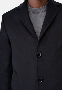 Svart ullblandad kappa med slagkrage, som har två framknappar och en ficka, tillsammans med en grå, ribbad rundhalsad tröja.