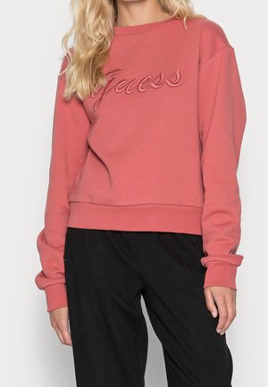 Vrouw draagt een koraalroze sweatshirt met "Guess" geborduurd op de borst en een zwarte broek, staand tegen een effen achtergrond.