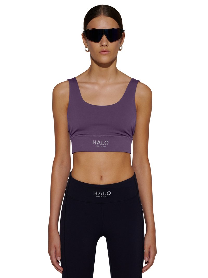 Kobieta nosząca fioletowy krótki top i czarne legginsy z logo "HALO", duże czarne okulary przeciwsłoneczne i srebrne kolczyki koła, stoi zwrócona przodem.