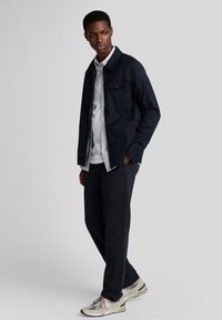 Marinblå jacka med knäppt ficka, vit skjorta med knappar, svarta byxor och ljusgrå sneakers. Avslappnad passform med släta texturer.