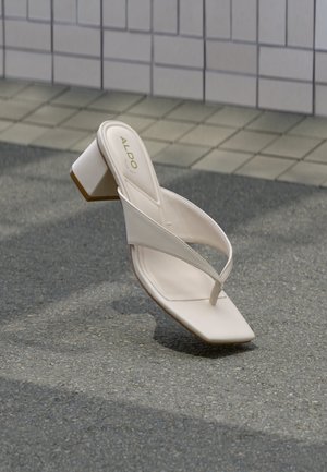 Sandale cu toc bej, cu vârf pătrat și barete încrucișate, pe asfalt gri, lângă un perete placat cu dale.