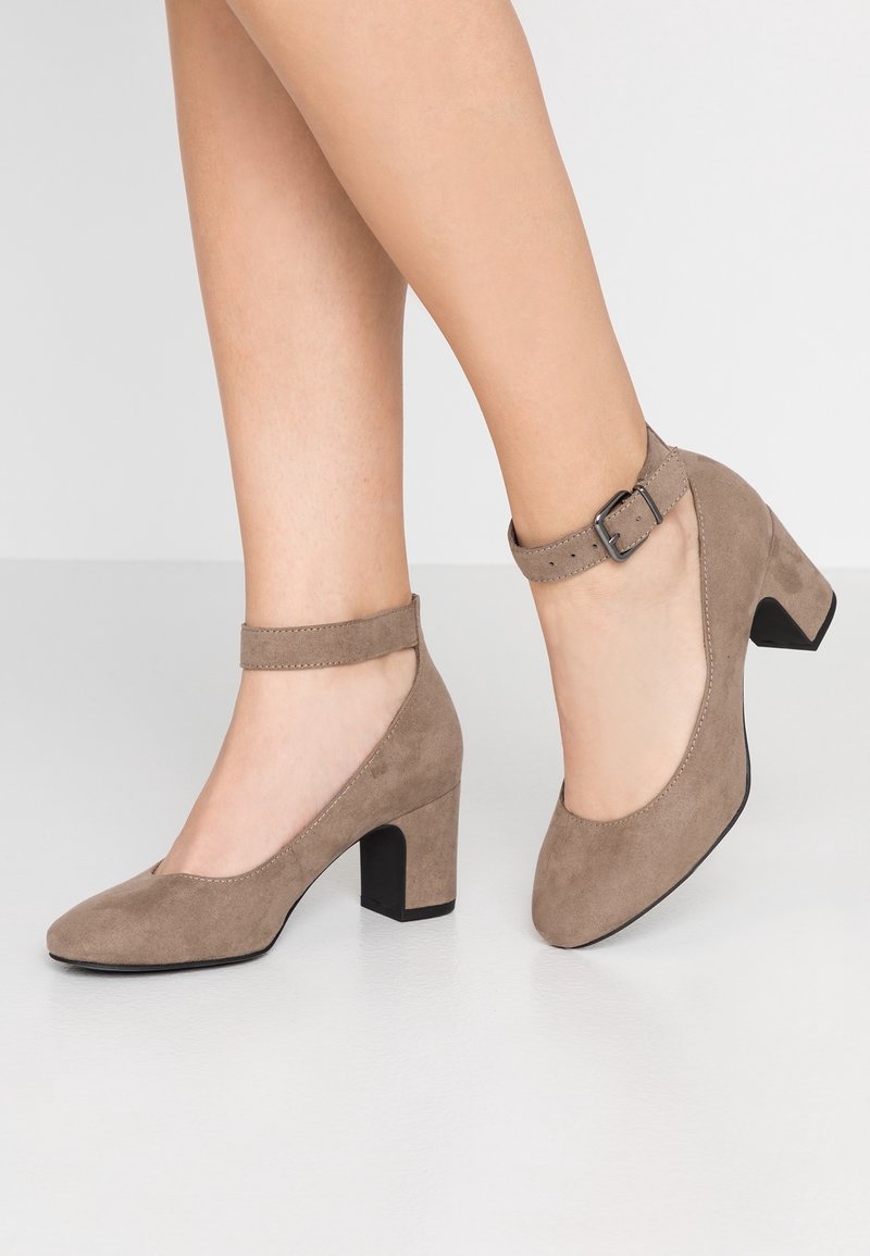 Chaussures à talons blocs en daim taupe avec brides à la cheville et boucles portées sur des jambes croisées sur un fond clair uni.