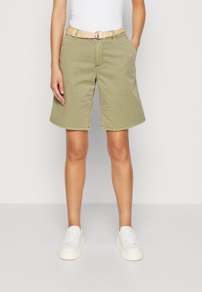 Personne portant un short vert olive jusqu'au genou avec une ceinture tissée beige, des baskets blanches et un haut blanc, debout, les mains détendues.