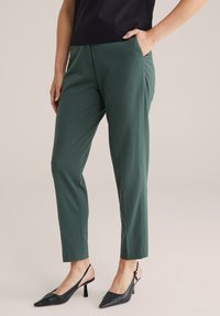 Žena oblečená ve zelených přizpůsobených kotníkových kalhotách, černých slingback podpatcích a černém topu, s jednou rukou v kapse, stojící na neutrálním pozadí.
