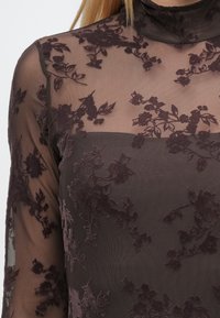 Blouse marron transparent avec broderies florales, col montant et manches longues. Tissu texturé avec des motifs de roses foncées sur toute la surface.