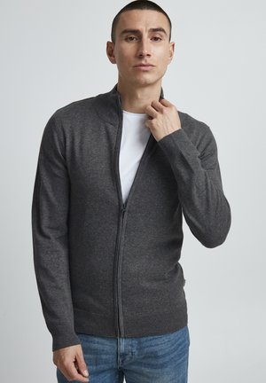 Mørk grå zip-up genser med høy krage, lange ermer, ribbestrikkede mansjetter og kant, brukt over en hvit t-skjorte. Jevn tekstur, minimalistisk design.