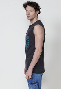 Svart ärmlös t-shirt med en blå grafisk tryck på framsidan, tillverkad av ett mjukt, texturerat tyg. Bärs tillsammans med blå cargoshorts.