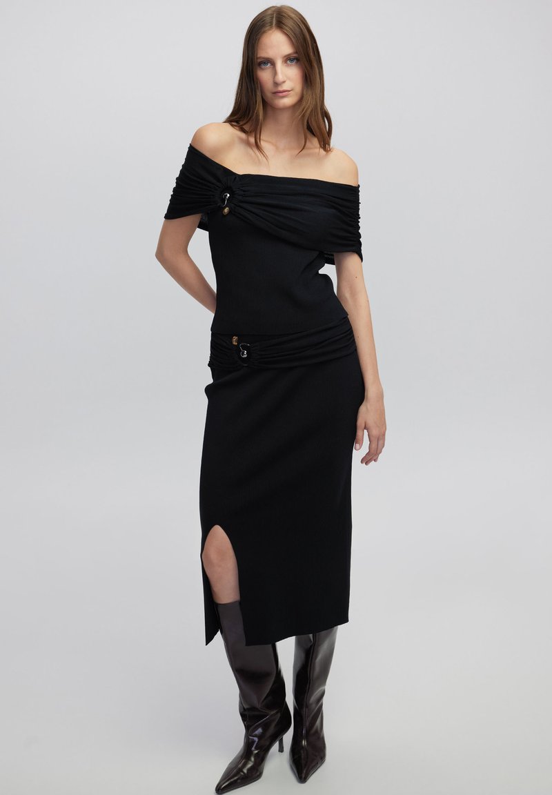 Abito nero off-the-shoulder con texture arricciata, spacco laterale e dettaglio con fibbia accentata in oro. Abbinato a stivali neri lucidi al ginocchio.