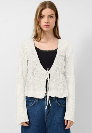 Junge Frau trägt eine weiße Strickjacke, die an der Taille gebunden ist, über einem schwarzen Top mit Spitzenbesatz und blauen Jeans vor einem neutralen Hintergrund.