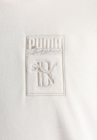 Tissu blanc avec un logo Puma en relief représentant un puma sautant et un texte stylisé à l'intérieur d'une bordure rectangulaire.