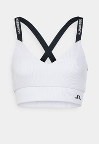 Bílá sportovní podprsenka s žebrovaným vzorem a černými překříženými ramínky, s logem na ramínku. Hladká textura a podpůrný pás na spodní části.