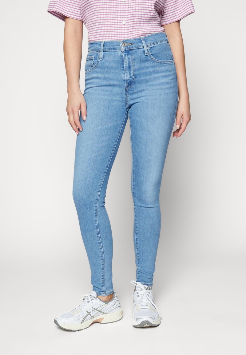 Jean skinny bleu clair en tissu denim avec une taille haute, doté de poches avant et d'un effet légèrement délavé. Porté avec des chaussures de sport blanches.