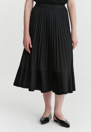 Personne portant une jupe midi plissée noire et des chaussures slingback noires à talon bas, debout devant un fond uni.