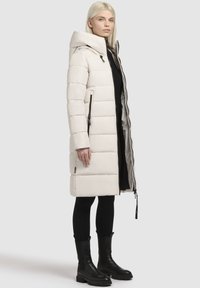 Manteau doudoune long et crème avec capuche, doté d'une fermeture éclair et de poches latérales. Design texturé et matelassé. Porté sur des leggings noirs et des bottes.