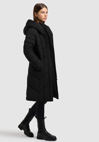 Lange zwarte pufferjas met een capuchon, voorzien van een gewatteerd patroon en zijzakken. Draagt met zwarte aansluitende leggings en stevige zwarte laarzen.