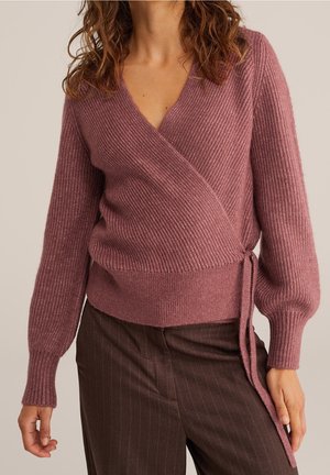 Femme portant un pull cache-cœur côtelé mauve avec de longues manches ballon nouées à la taille, associé à un pantalon marron foncé à fines rayures.