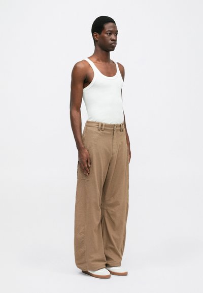 Bēša krāsas vaļīgas kravas bikses ar sānu kabatām un jostu viduklī. Kombinētas ar piegulošu baltu topiņu un gaiši krāsotiem apaviem.