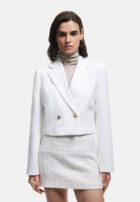 Hvid dobbeltradet jakke med tekstureret stof, guldknapper og et cropped design; parret med en matchende miniskørt i lignende materiale.