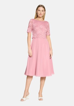 Woman wearing a pink lace top dress with elbow-length sleeves and a flowy skirt, paired with white open-toe wedge sandals, standing on white background.