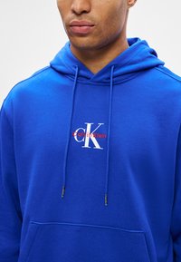 Férfi világoskék Calvin Klein kapucnis pulóvert visel, fehér és piros logóval, amely a mellkastól az állig látható, fehér háttér előtt.