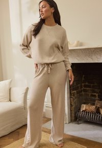 Beige ribbattuja neuleita oleva loungewear-setti, jossa on pyöreä pääntie, nyöritettävät, leveät housut, rento istuvuus ja pehmeä rakenne.