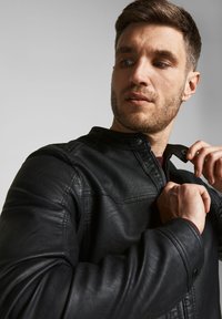 Veste en cuir noir avec un col haut, dotée d'une fermeture éclair et de boutons-pression. Texture douce et design ajusté avec des détails de couture visibles.