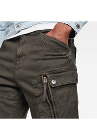 Oliivivihreät cargo-housut, joissa on kulunut pinta, vinot etutaskut ja vetoketjullinen sivutasku, jossa on metalliset viimeistelydet. 