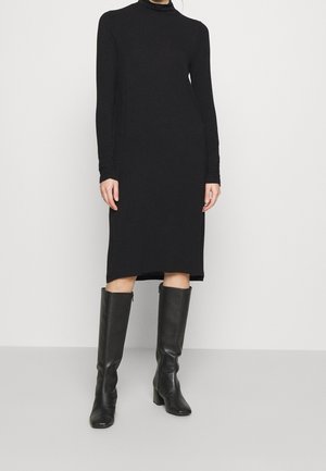 Frau trägt ein schwarzes Midi-Kleid mit langen Ärmeln und kniehohe schwarze Lederstiefel, steht vor einem schlichten hellen Hintergrund.