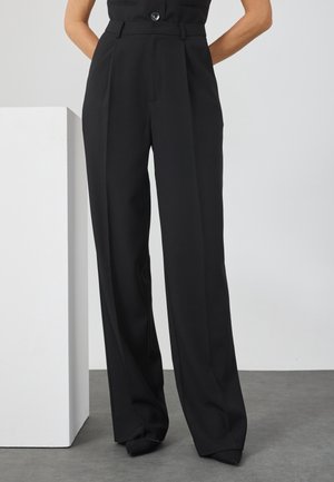 Kvinde iført højtaljede, brede sorte bukser med læg, står med hænderne gemt bag ryggen mod en ensfarvet baggrund.